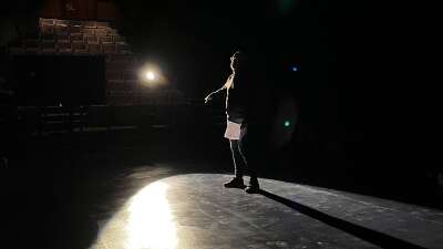 A man appears under a spotlight on PTE's empty stage. He is backlit and appears mostly in silhouette. In his hand, he is holding a script.